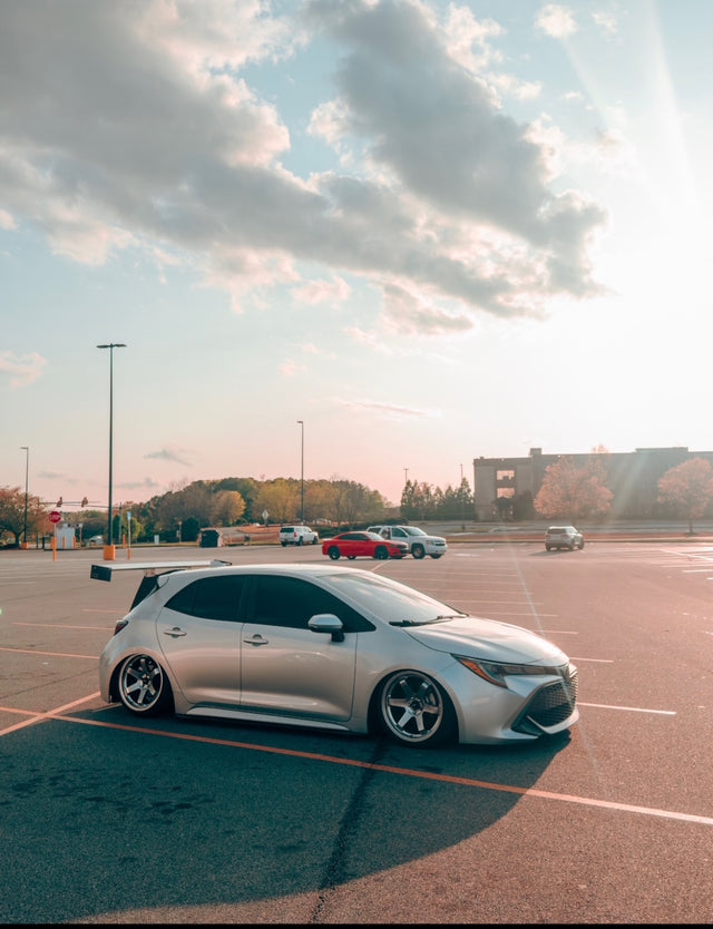 White Toyota Corolla E210 in an open parking lot under a colorful evening sky.