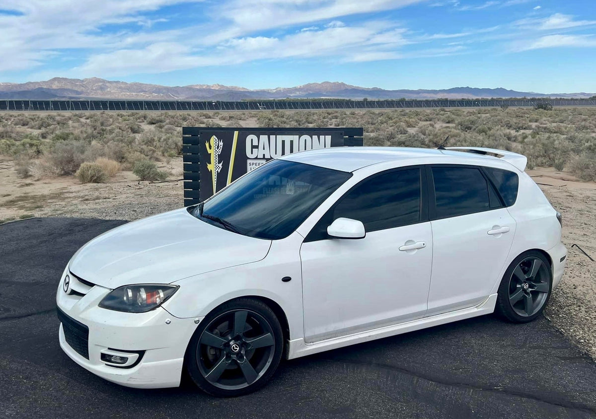 White first-generation Mazdaspeed3 with black wheels, shown from the front quarter outdoors.