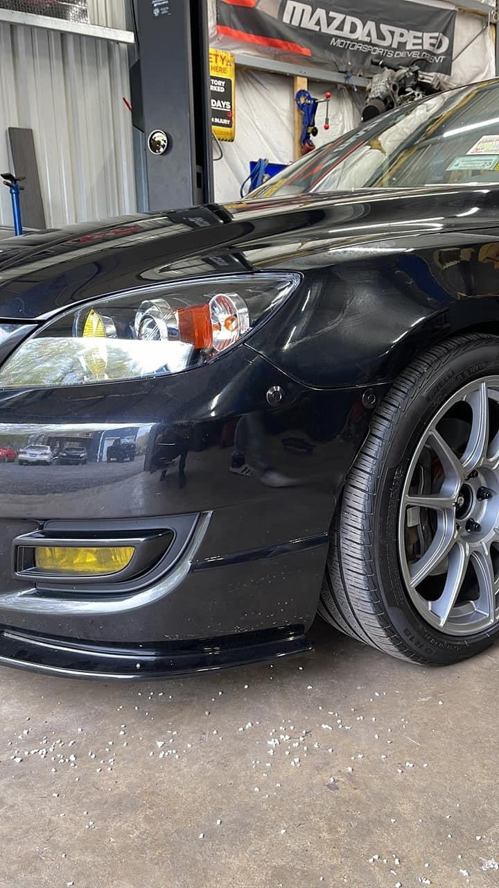 Black first-generation Mazdaspeed3 showing two dark latches along the front bumper-to-fender seam.