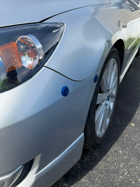 Closeup of two blue bumper latches fitted below the headlamp and beside the wheel of a silver Mazda3.