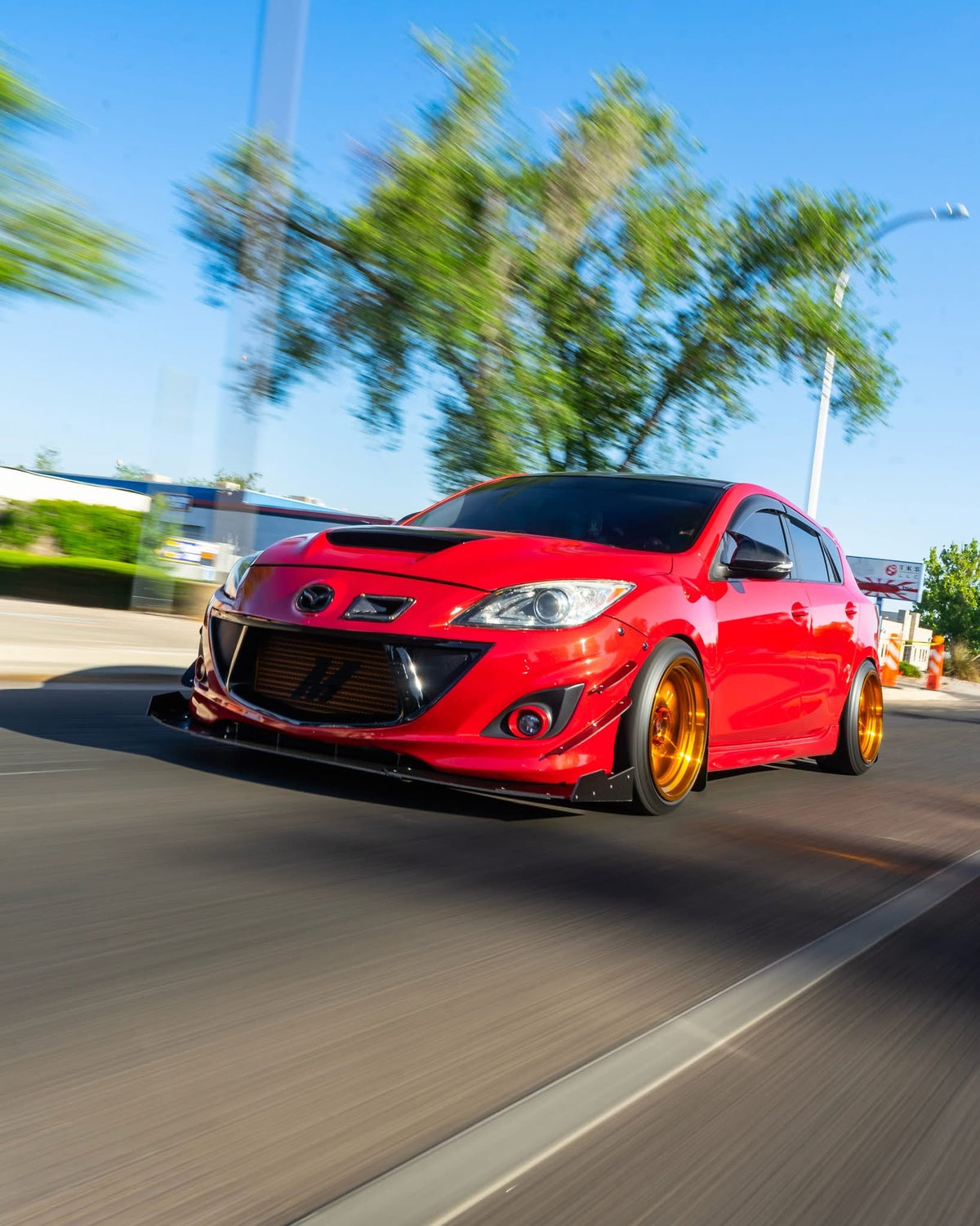 Red second-generation Mazdaspeed3 with gold wheels, viewed from the front quarter on the road.