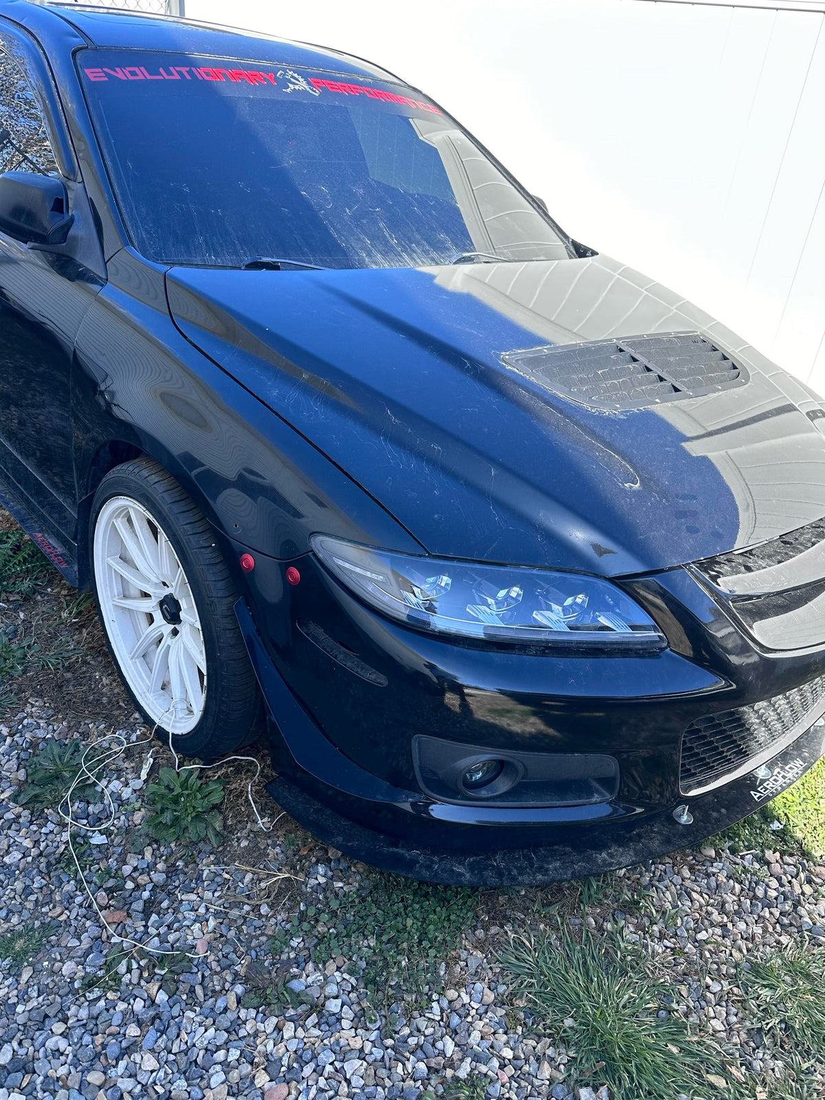 Two red bumper latches installed beside the front wheel of a dark Mazda6 with white wheels.