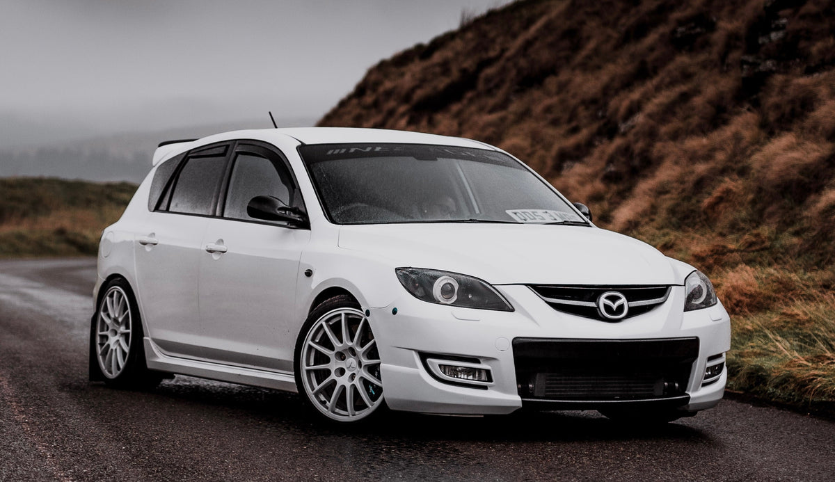 White first-generation Mazdaspeed3 with front bumper latches, photographed beside a hillside.