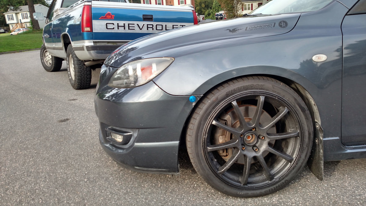 Blue latch installed at the front wheel arch of a gray first-generation Mazda3.
