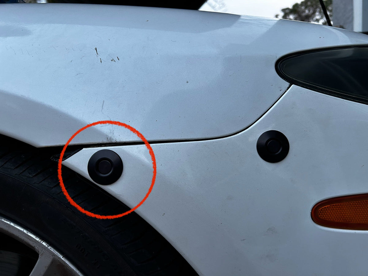 Red circle marking the wheel-arch bumper latch on a white car, with a second latch near the headlamp.