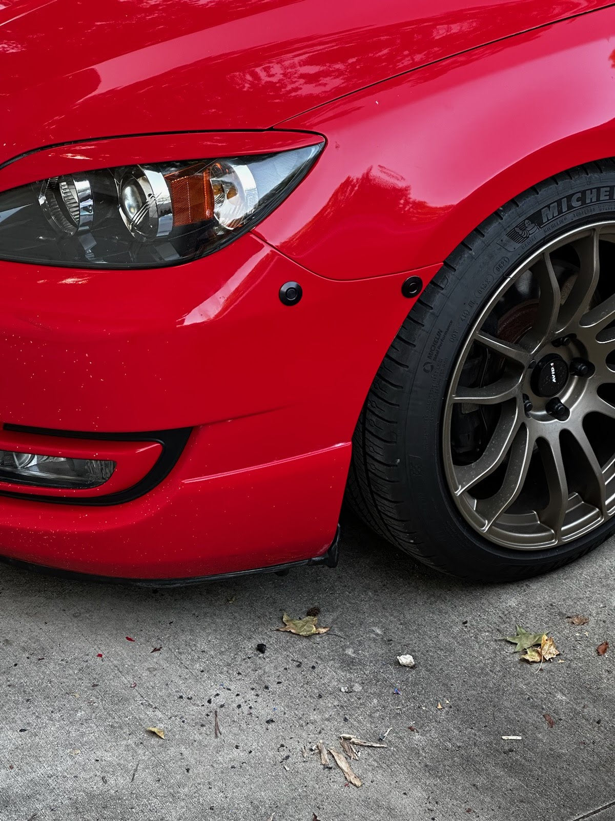 Two black bumper latches installed below the headlamp and beside the wheel of a red Mazdaspeed3.